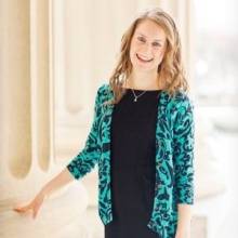 Maura is standing in front of a cream colored stone pillar. Her right hand is resting on the pillar. She is wearing a black dress with a teal colored cardigan with black swirls.