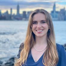 Maggie is standing in front of a river or lake. There is a city on the other side of the water. She is wearing a blue shirt with a V-neck.